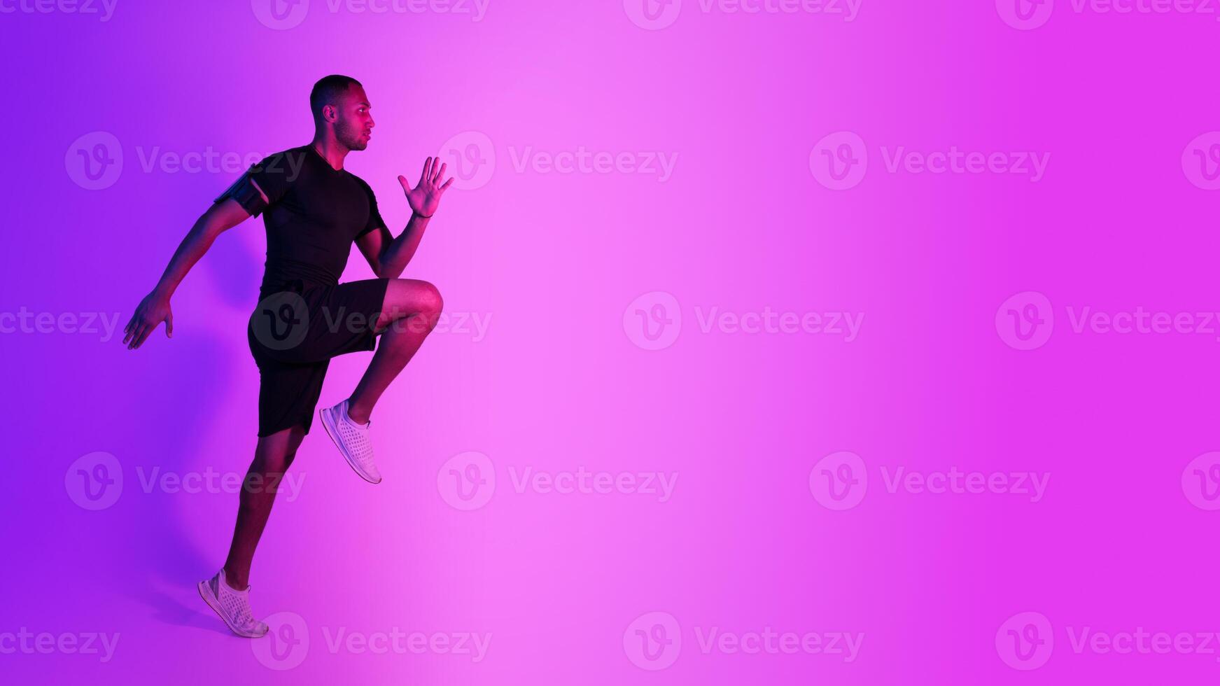 Black Athlete Guy Jumping Exercising During Training On Purple Neon Studio Background, Looking Aside At Copy Space. Full Length, Side View Shot. Male Fitness Workout And Sport Motivation. Panorama photo