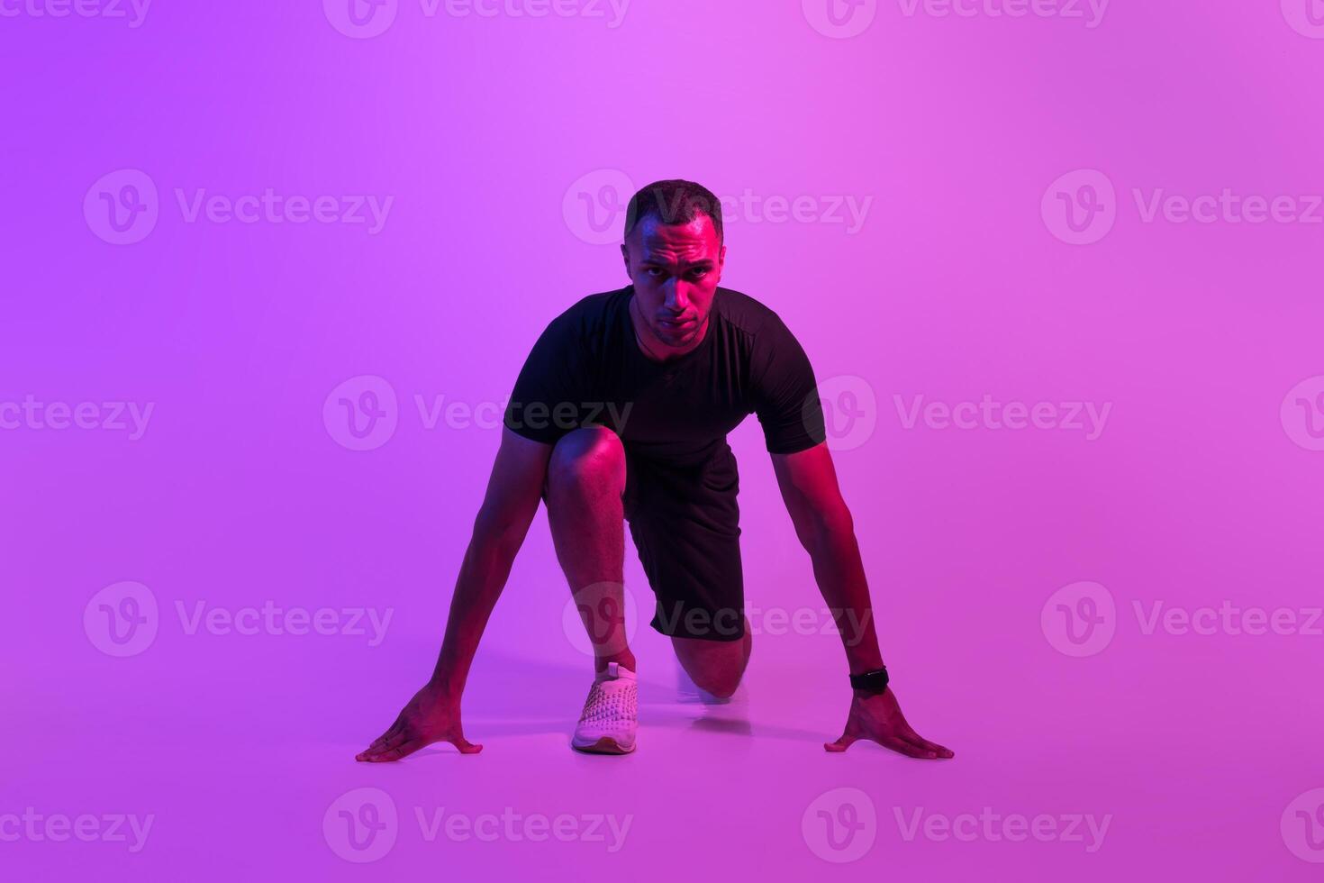 Front View Of Motivated African American Runner Guy Doing Crouch Start And Looking At Camera, Sportsman Posing Ready For Race On Purple Neon Background. Fit Man Preparing For Running Workout photo