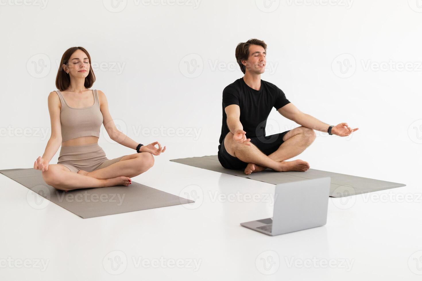 Positive calm millennial caucasian couple in sportswear training, practicing yoga with laptop on lotus position in gym, on white background. Workout remote, health care, lifestyle, sports and fit photo
