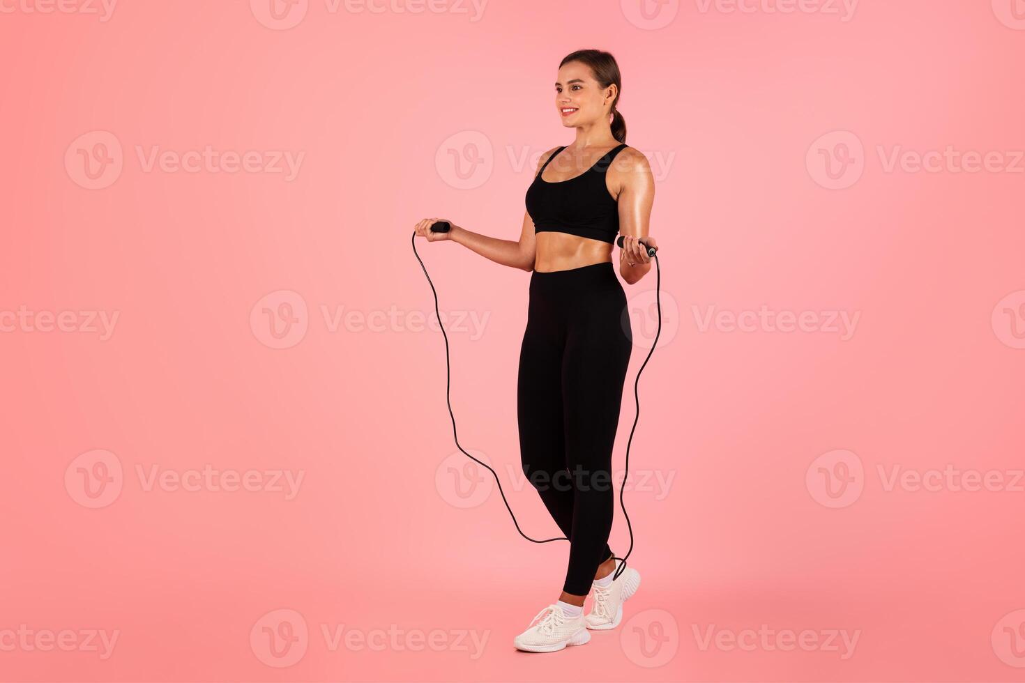Cardio Workout. Portrait Of Sporty Young Female In Activewear Holding Skipping Rope, Smiling Fit Woman Using Sport Equipment While Posing At Pink Studio Background, Looking At Copy Space, Full Length photo