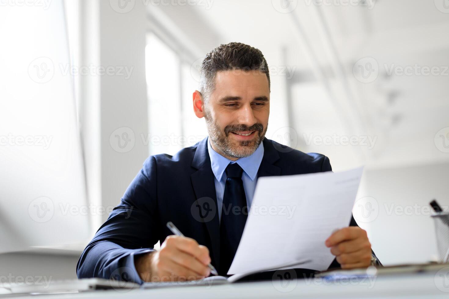 Positive middle aged businessman working with papers at workplace in office, entrepreneur man reading financial reports and taking notes, enjoying company growth and smiling photo