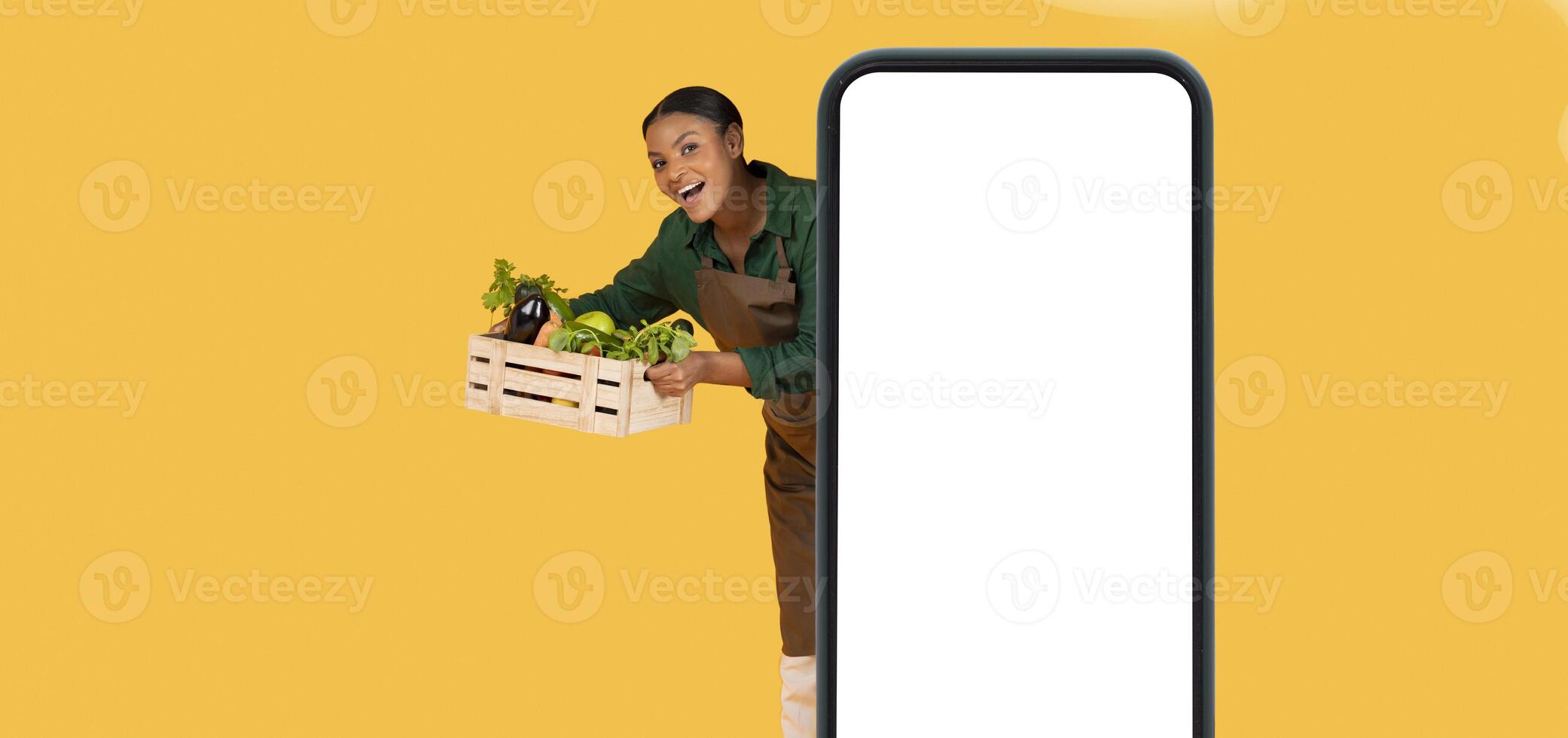 Grocery Market Application. Joyful Black Farmer Woman Posing Behind Large Smartphone Empty Screen, Holding Box With Fresh Vegetables, Showing Harvest From Garden, Yellow Background, Panorama photo