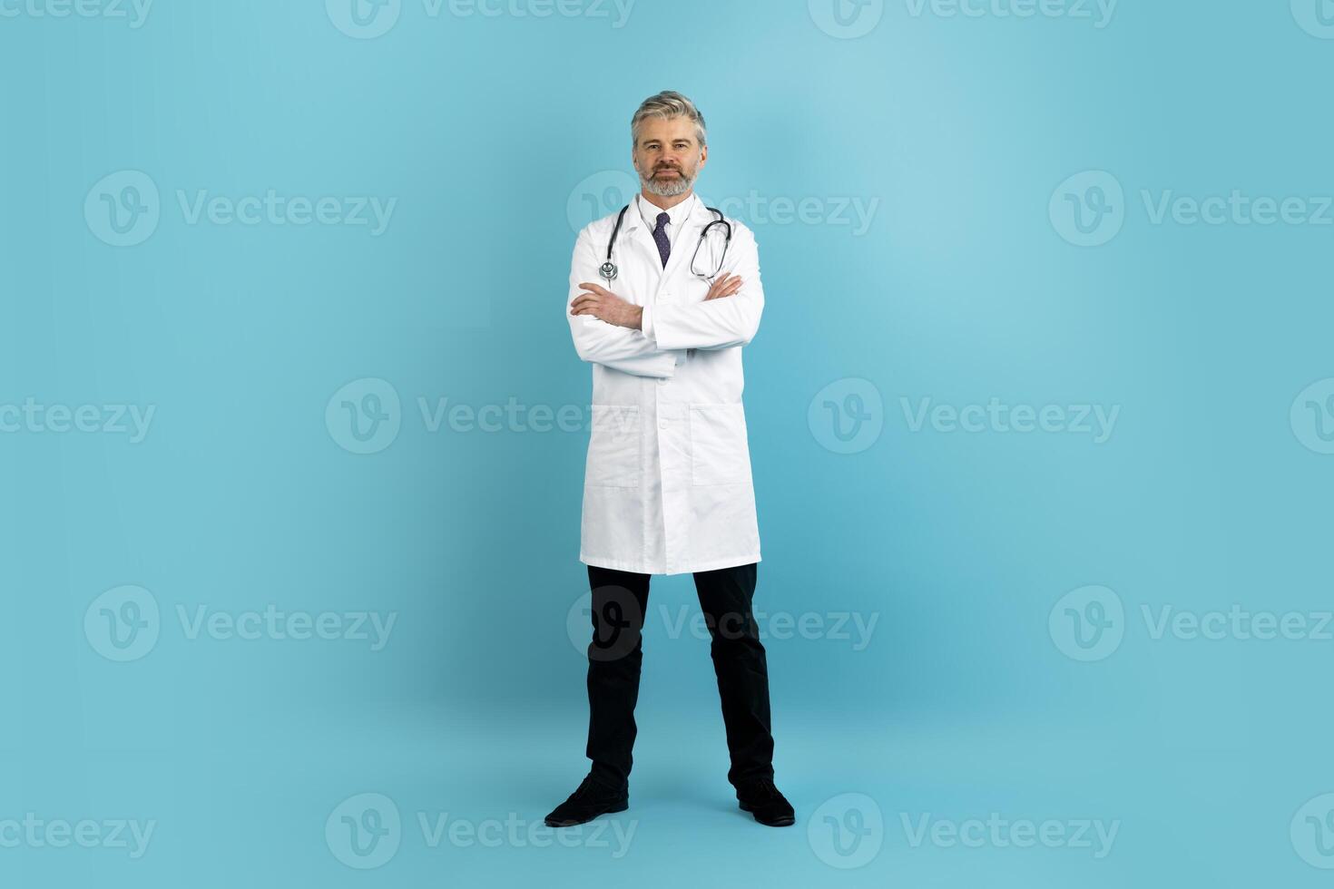 Serious handsome grey-haired bearded middle aged man doctor in medical white coat with stethoscope on his shoulders posing on blue studio background, holding arms crossed on chest, copy space photo