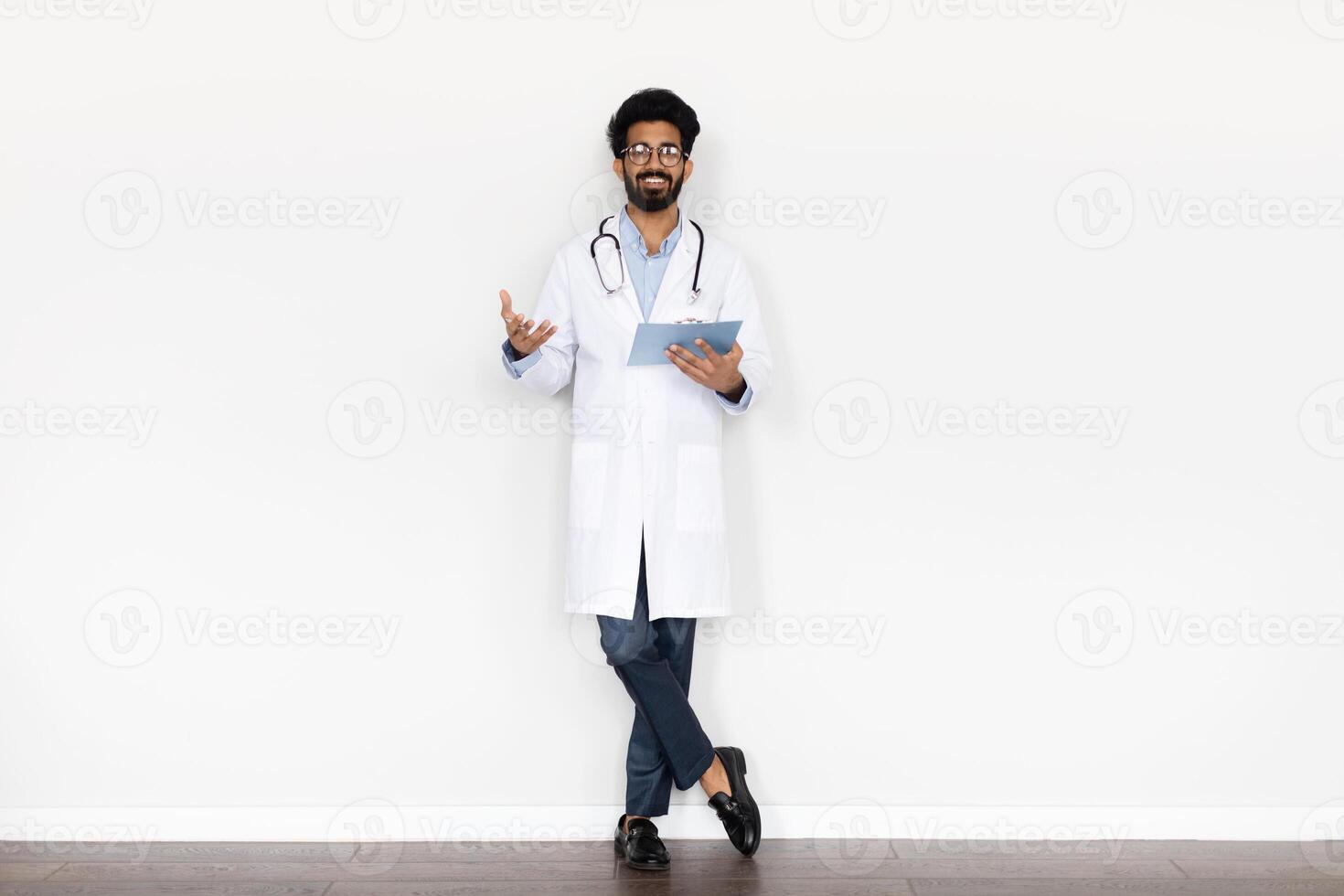 Friendly smiling handsome bearded young middle eastern doctor in medical coat with stethoscope and medical chart posing on white background, getting anamnesis, copy space. Healthcare, medical staff photo