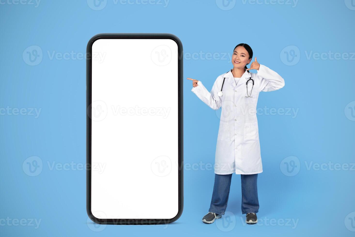 Smiling Asian Doctor Lady Gesturing Call Me While Standing Near Big Blank Smartphone, Therapist Woman Pointing At Mobile Phone With White Empty Screen, Posing On Blue Studio Background, Mockup photo