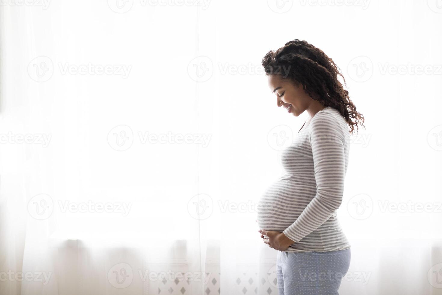 Profile Portrait Of Black Smiling Pregnant Woman Tenderly Embracing Belly While Standing Near Window At Home, Happy Expectant Mother Enjoying Her Healthy Pregnancy And Awaiting Time, Copy Space photo