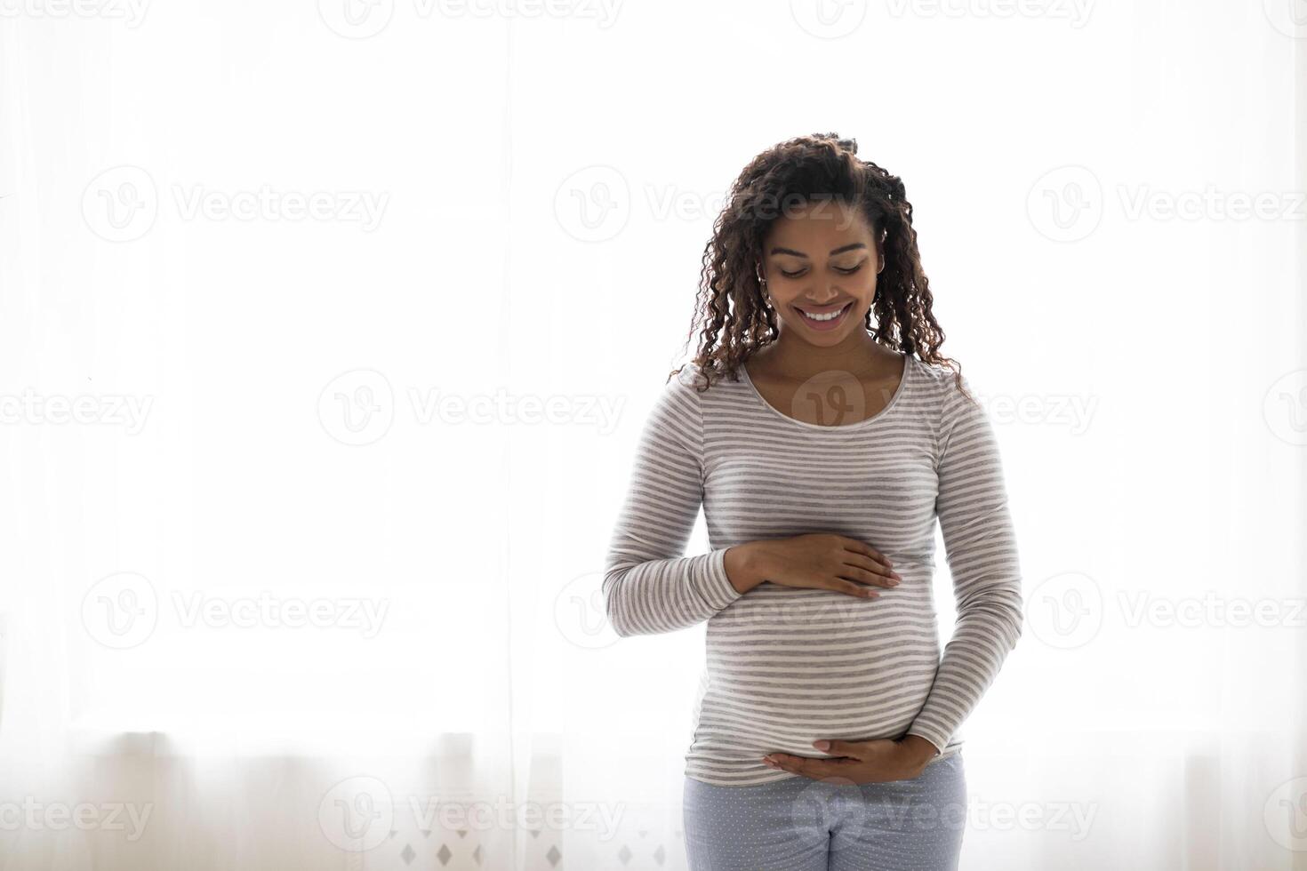 Portrait Of Beautiful Black Pregnant Woman Standing Near Window At Home, Embracing Belly. Young African American Expectant Mother Tenderly Touching Tummy And Smiling, Enjoying Pregnancy, Copy Space photo