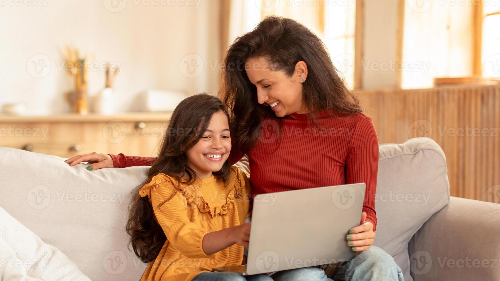 Joyful Middle Eastern Mom And Schoolgirl Daughter Using Laptop At Modern Living Room, Sitting With Computer, Browsing Internet And Having Fun Online At Home On Weekend. Panorama photo