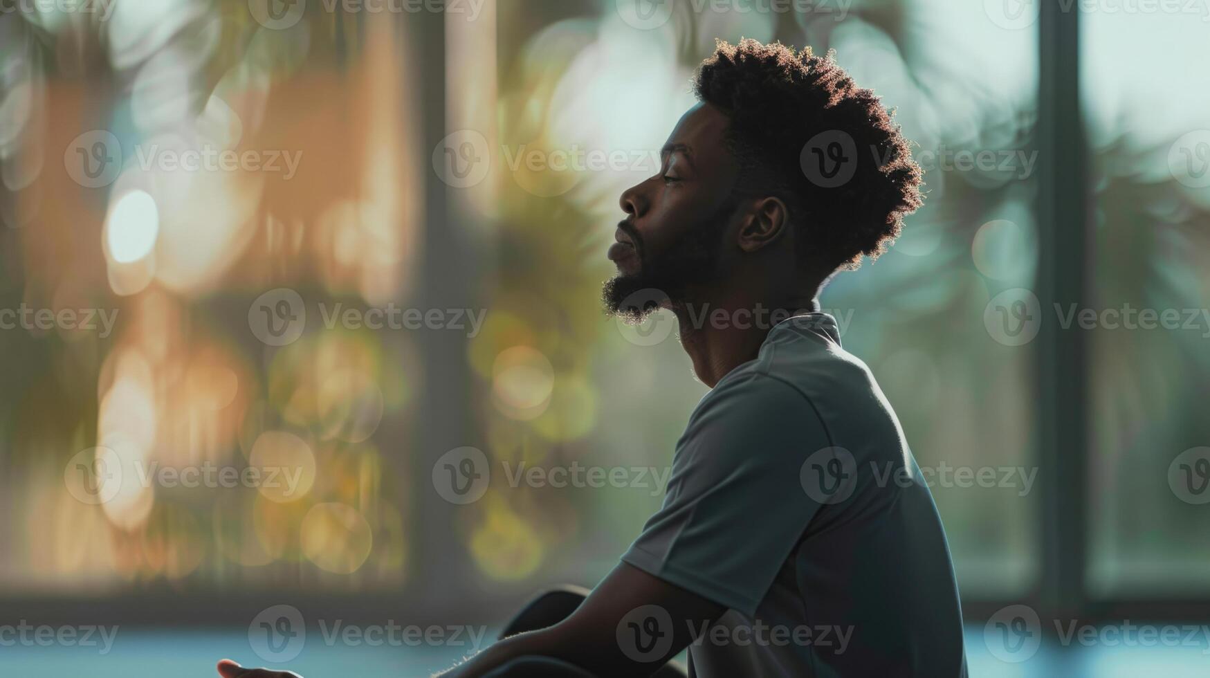 A man sits with his eyes closed in a meditative pose, bathed in soft afternoon light near a large window. photo