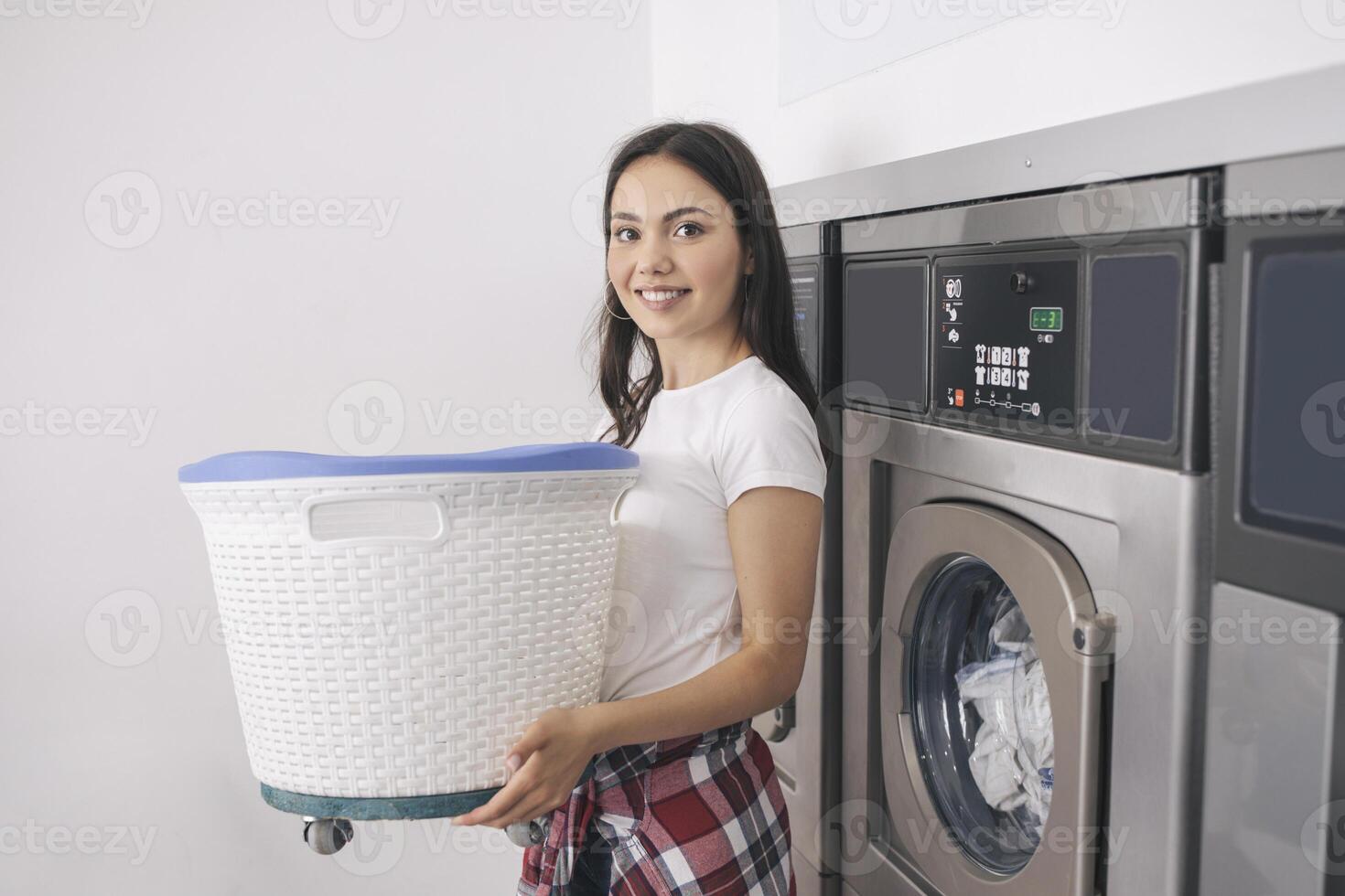 Laundromat Room. Happy Young Lady In Casual Doing Laundry At Public Laundrette Indoors, Posing Holding Basket With Dirty Clothes Near Industrial Washing Machines And Smiling To Camera photo