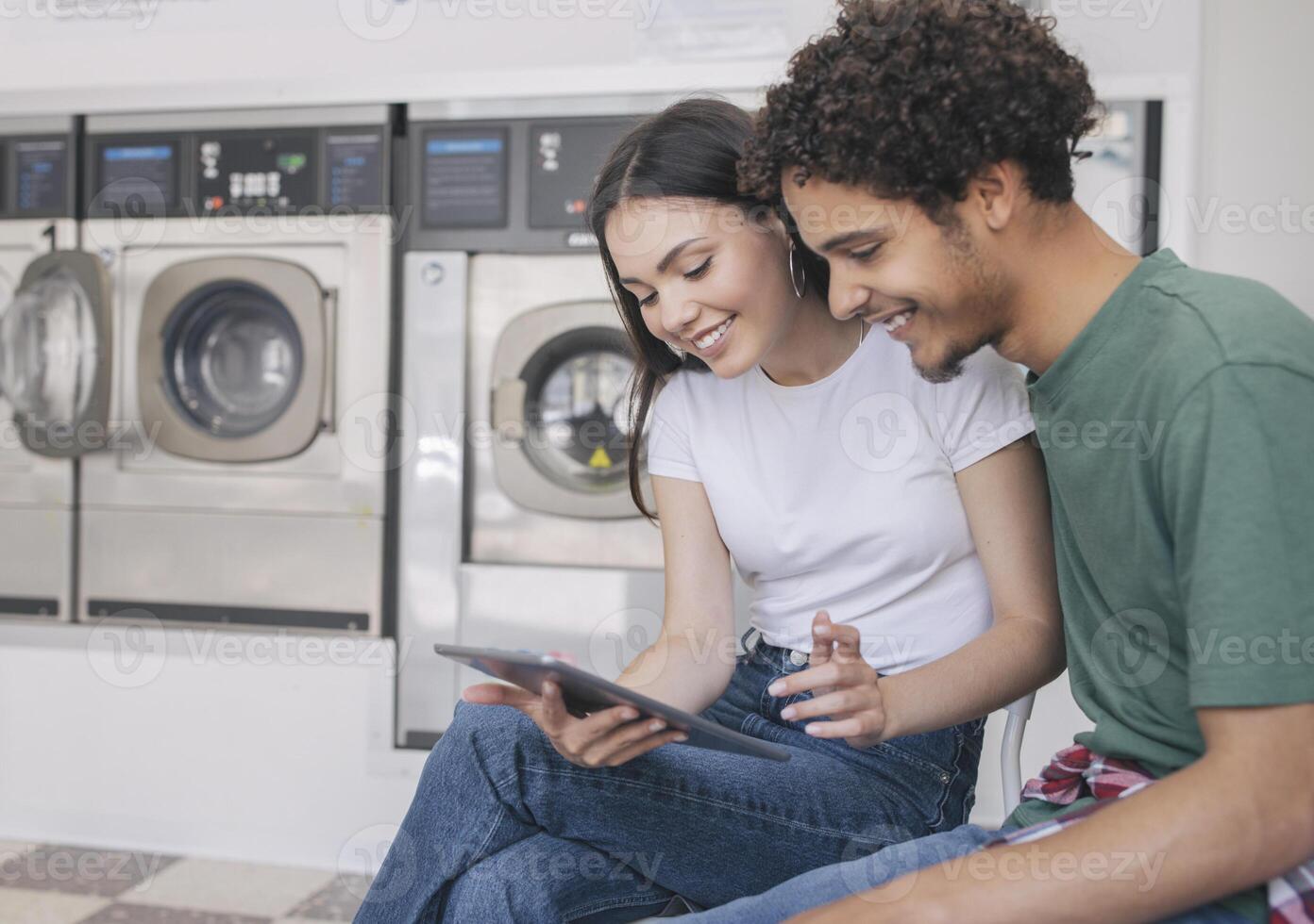 Happy Young Hispanic Couple Using Digital Tablet While Doing Laundry At Modern Laundromat Service Indoors. Man And Woman With Computer Waiting For Clean Clothes At Laundrette. Selective Focus photo