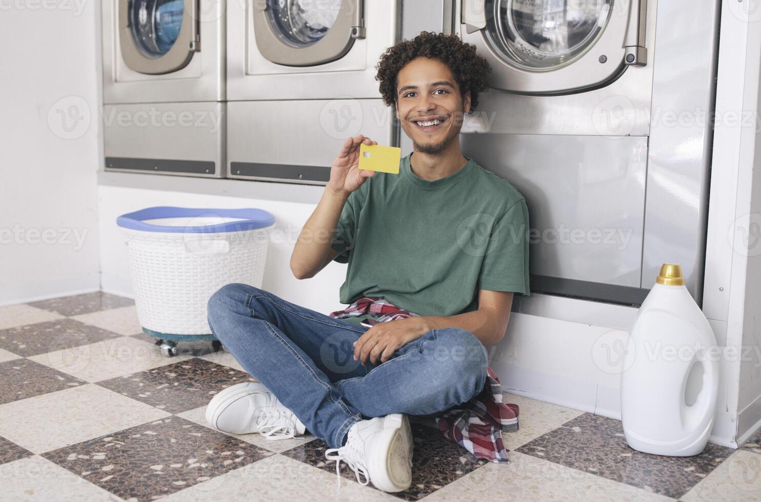 Young Latin Man Showing Credit Card Sitting With Detergent Bottle At Laundry Room Indoor, Smiling At Camera Posing Near Row Of Industrial Washing Machines. Great Public Conveniences Offer photo