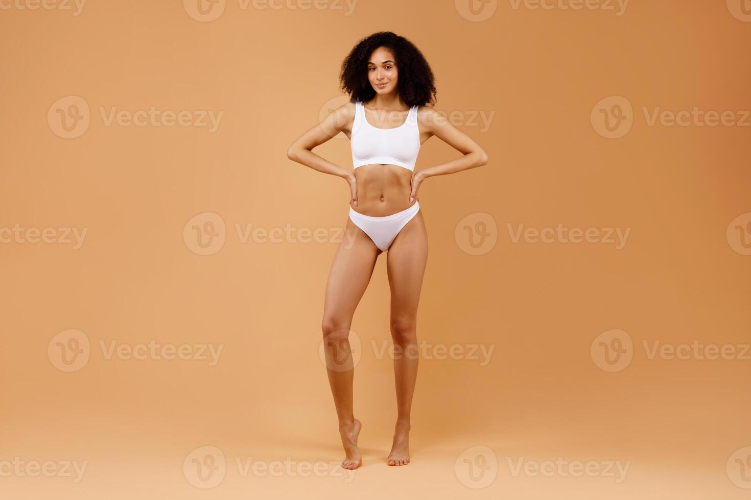 Full length shot of young beautiful latin lady posing in white underwear bra and panties over beige studio background, holding hands on her waist, demonstrating nice curves photo