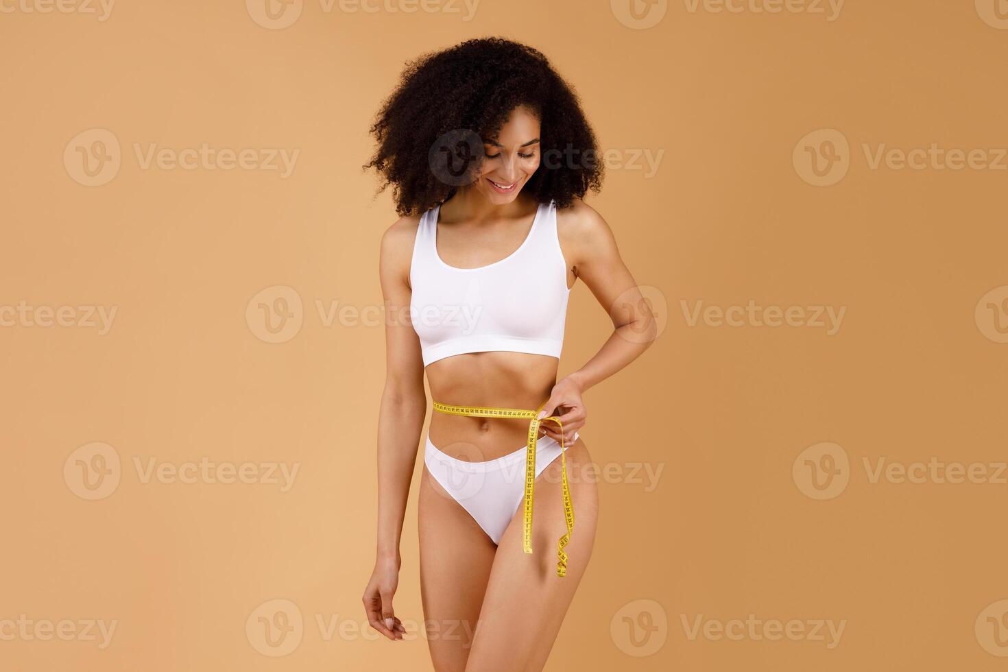 Satisfied happy well-fit black lady wearing white underwear holding measuring tape around her waist and smiling, posing isolated over beige background, copy space photo