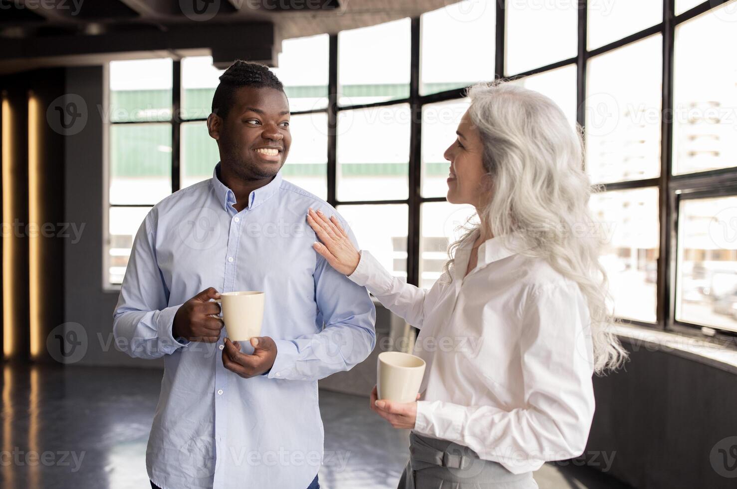 Corporate Lifestyle And Communication. Multiethnic Business Colleagues Enjoying Coffee Break, Talking Holding Mugs, Lady Touching Man's Shoulder Communicating In Office Interior photo