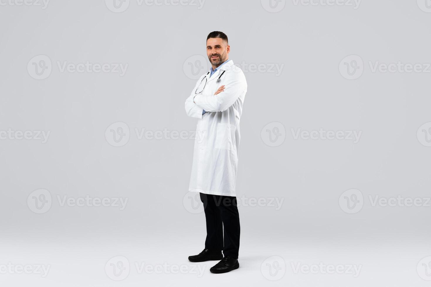 Cheerful middle aged man doctor posing with folded arms looking at camera standing on light background, wearing white uniform, coat and stethoscope, full body length, free space photo