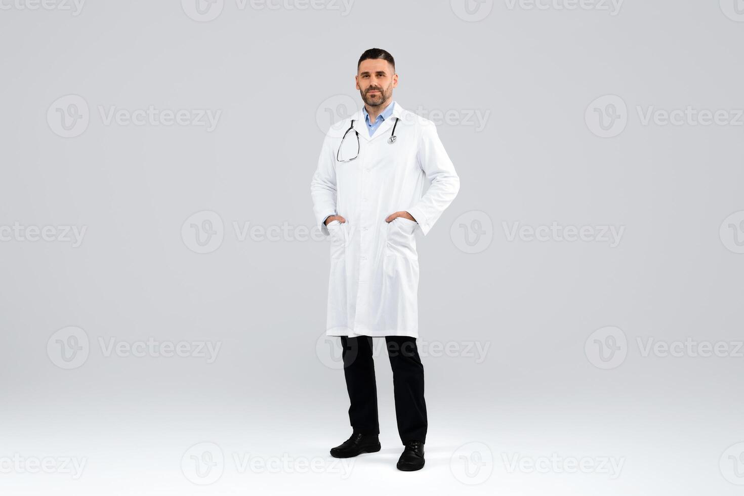 Confident middle aged doctor in white workwear looking at camera, posing with hands in pockets on light studio background, full length. Healthcare, medical, clinic concept photo