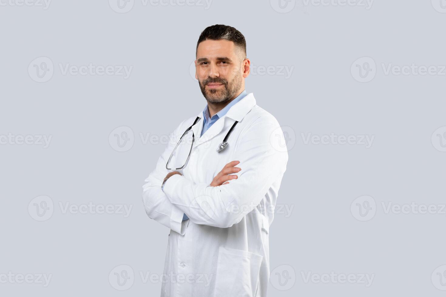 Medical services concept. Portrait of middle aged man doctor therapist in uniform posing with folded arms and looking at camera over light studio background, copy space photo