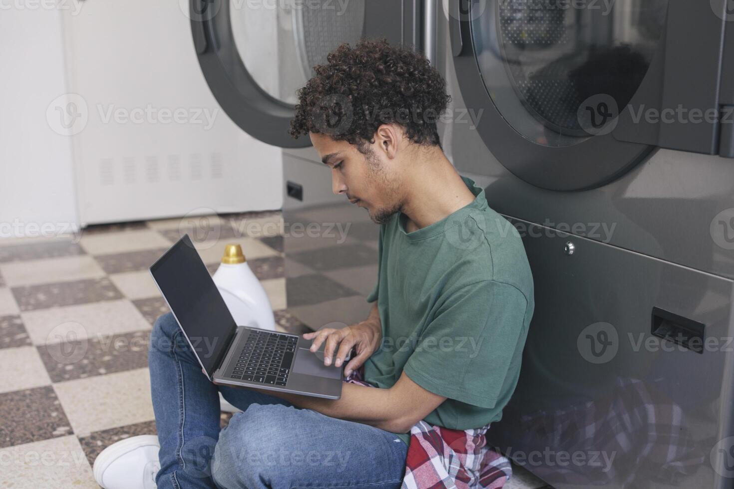 Great Laundry Service. Middle Eastern Student Guy Learning Online Via Laptop While Washing Clothes At Public Laundrette, Sitting Indoor With Computer And Surfing Web, Wearing Casual. Side View photo