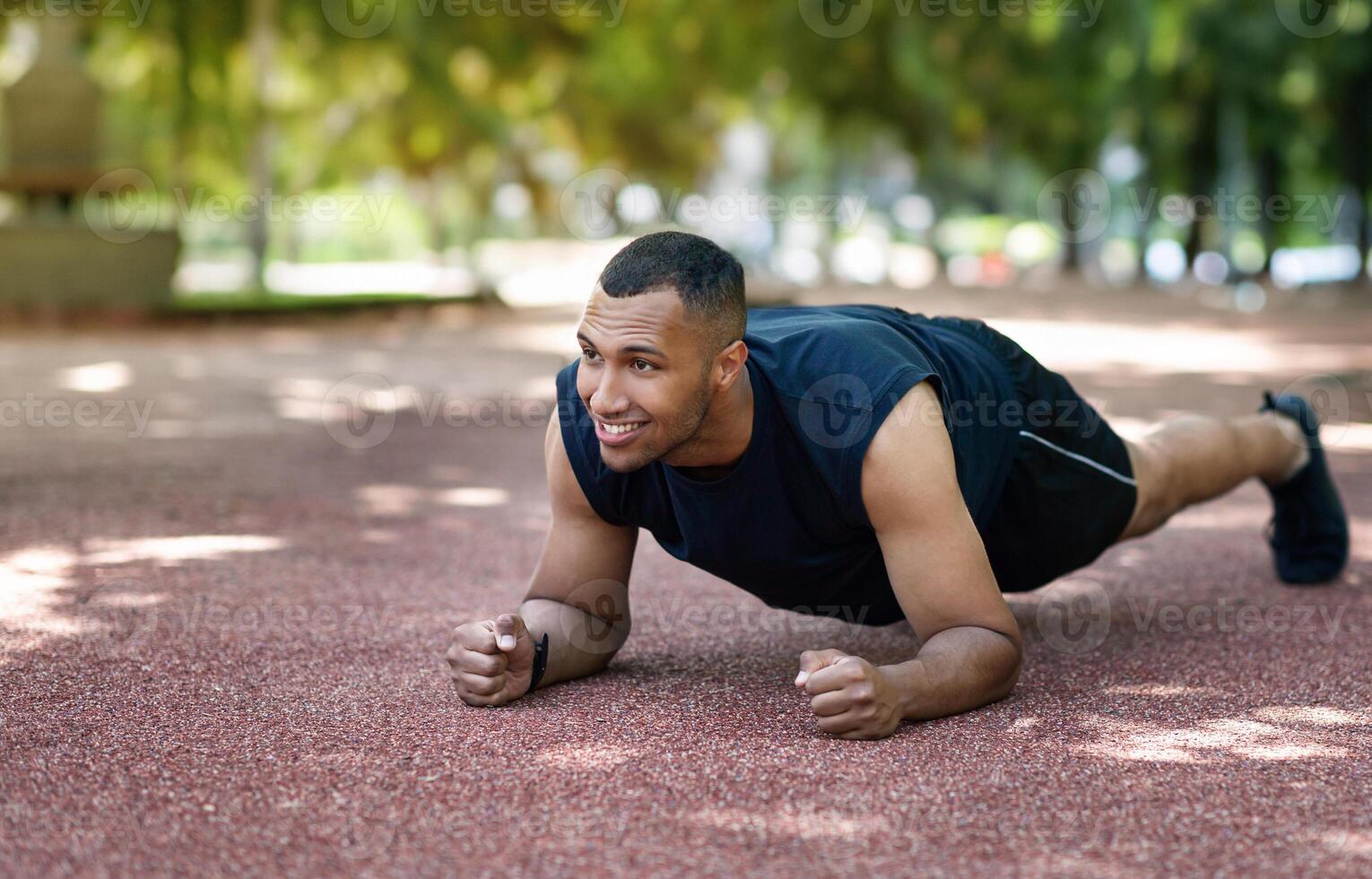 Positive strong african american guy standing in elbow plank on road at park at morning, panorama. Workout for muscles, power and motivation in city, sports outdoor, active lifestyle photo