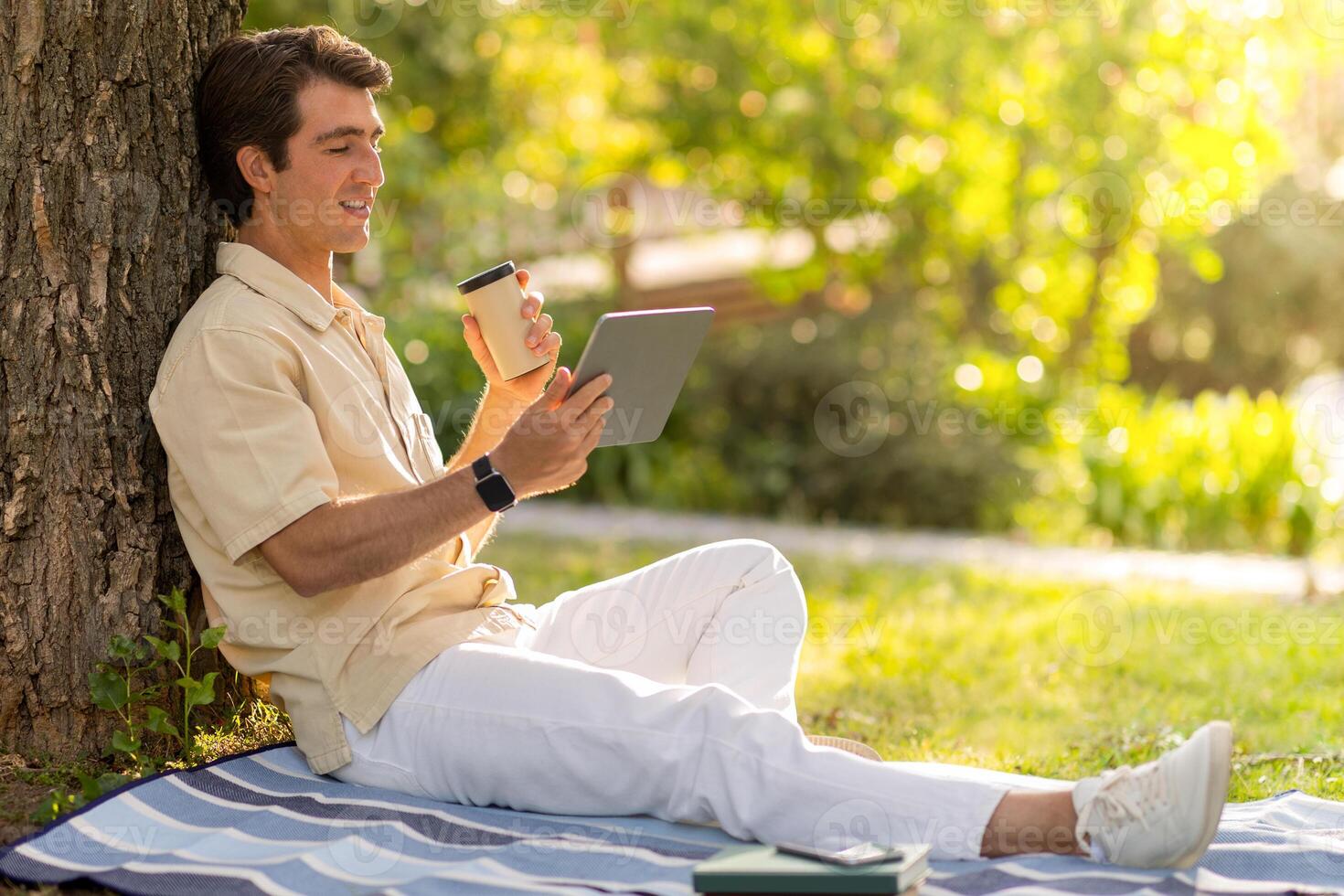 Happy handsome guy wearing casual outfit sitting under tree at public park, using digital tablet, copy space. Millennial man resting outdoors, have call with family or lover. Telecommunication photo