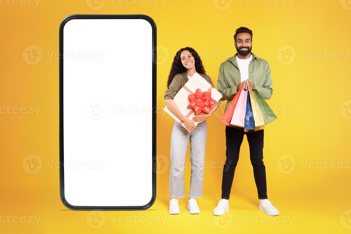 Shopping App. Cheerful Millennial Arab Customers Couple Near Big Cellphone With Empty Screen On Yellow Studio Background, Holding Gift Box And Colorful Shopper Bags. Mockup, Sales Offer Ad. Collage photo