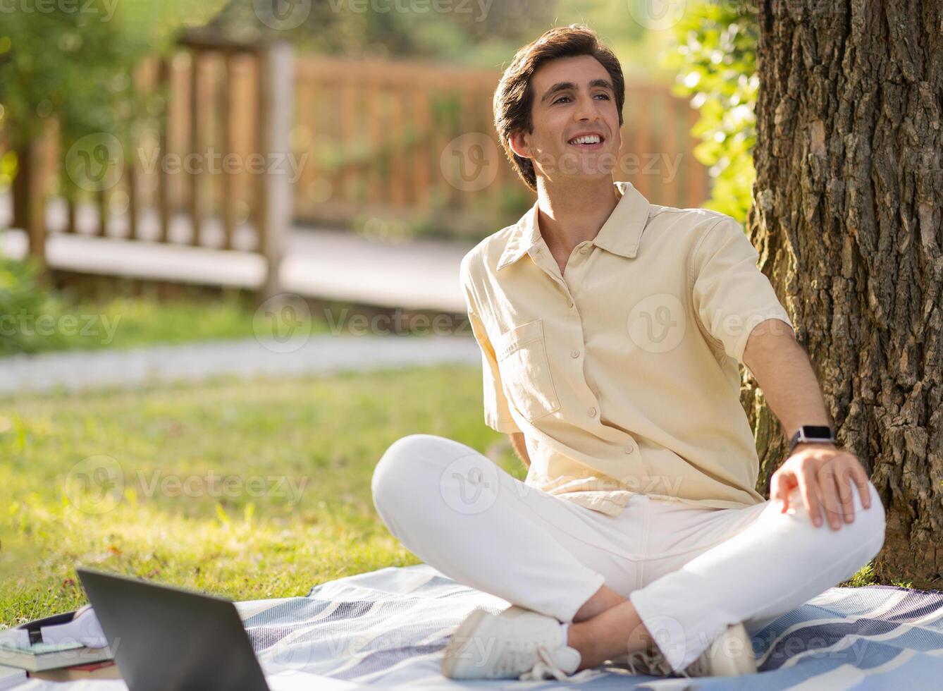 Positive attractive caucasian young man wearing casual outfit sitting on plain blanket under tree at park, have picnic, working online, using laptop, enjoying sunny summer day, looking at copy space photo