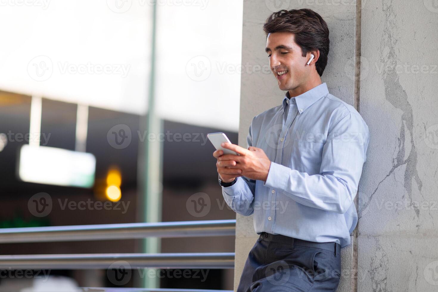Positive handsome young man in fromal outfit employee standing at parking place next to office building, using smartphone and wireless earphones outdoors, watching content, copy space photo