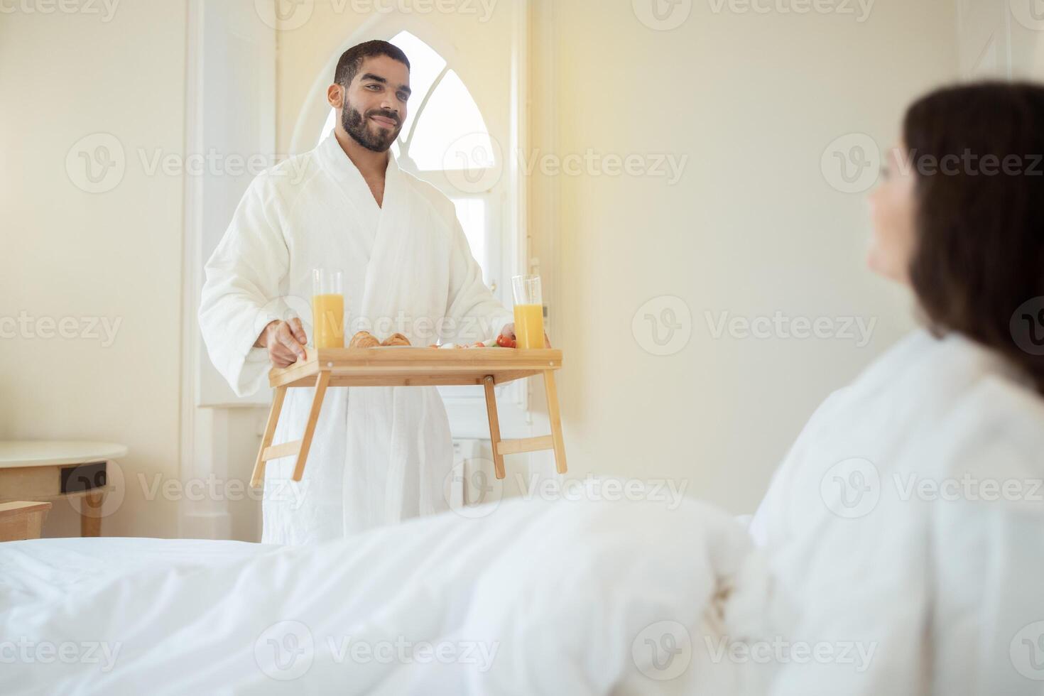 Honeymoon Morning. Husband Delivering Gourmet Breakfast Tray To Wife Celebrating Romantic Vacation, Relaxing In Bed In Luxurious Hotel Suite Indoors. Romantic Gestures And Relationship photo