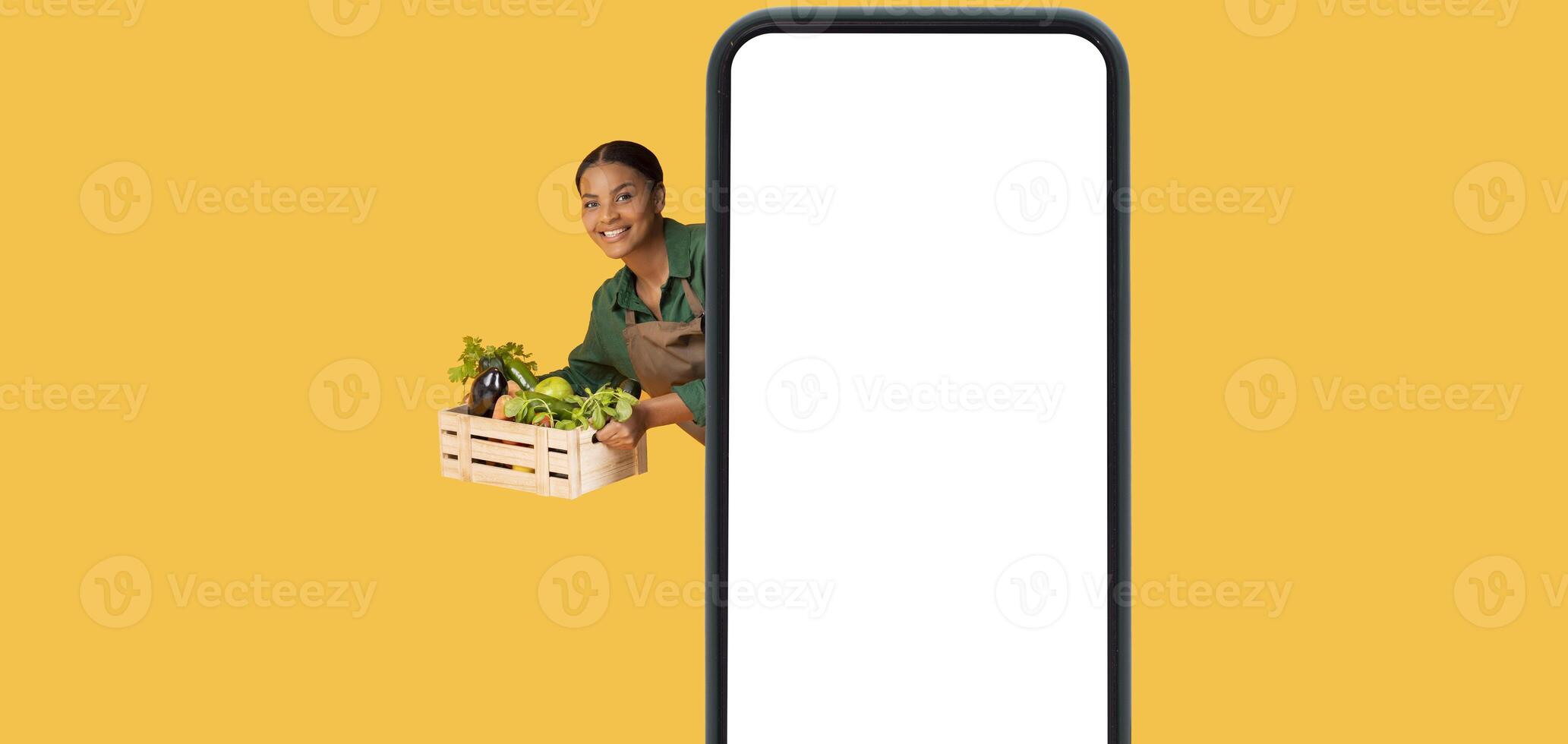 Online Farm Business. Collage With Big Phone Blank Screen And African American Farmer Lady With Box Of Garden Vegetables, Posing Behind Cellphone And Showing Harvest On Yellow Background. Panorama photo