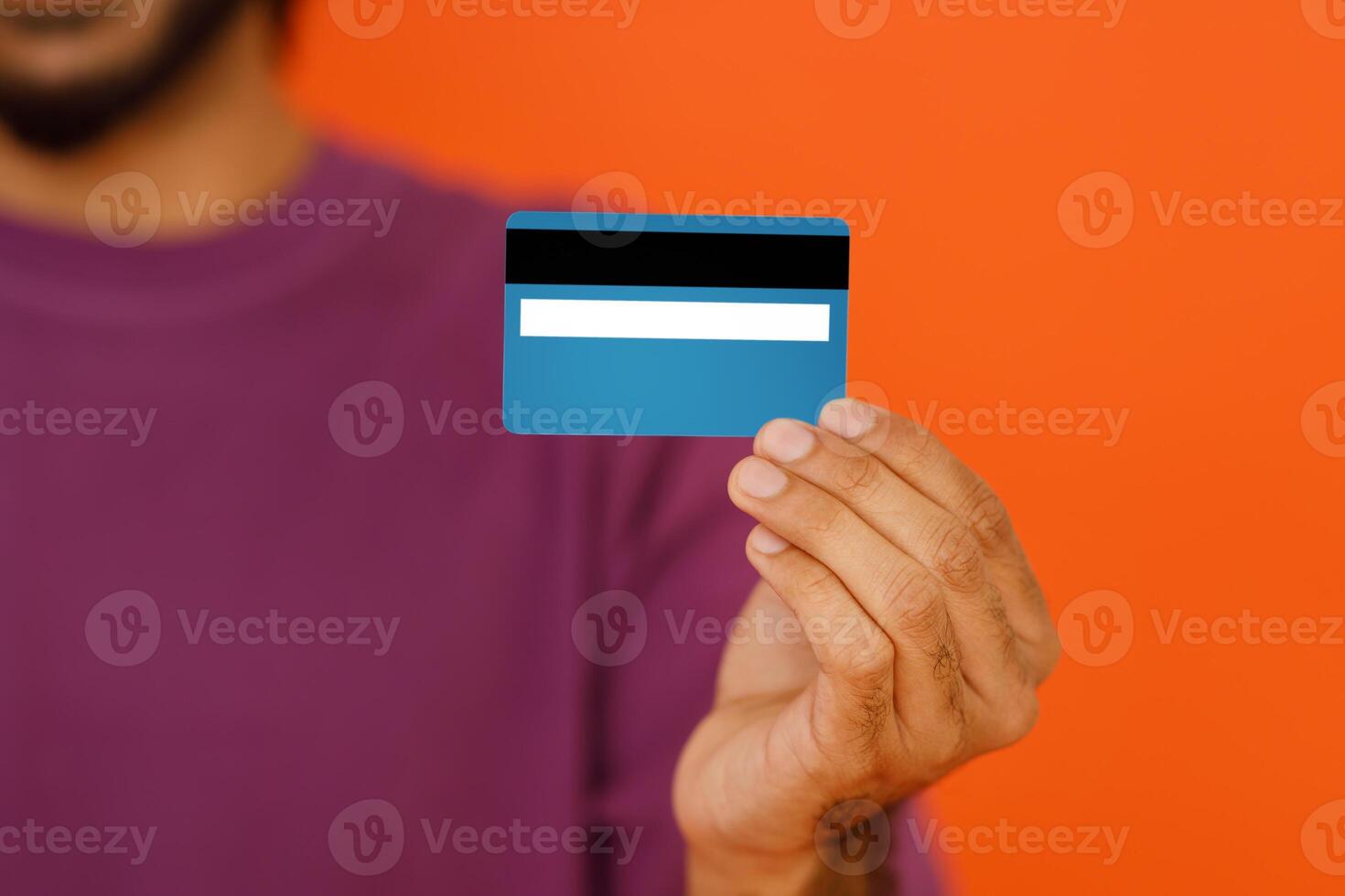 Banking concept. Cropped image of young dark-skinned man wearing basic casual t-shirt holding in hand credit bank card isolated on bright orange colour background, studio shot, blurred, copy space photo