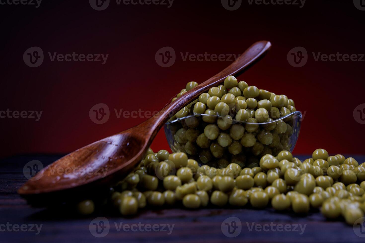 A glass cup with green canned peas stands on a wooden surface. A wooden spoon lies on a cup with canned green peas. Beautifully illuminated background. photo