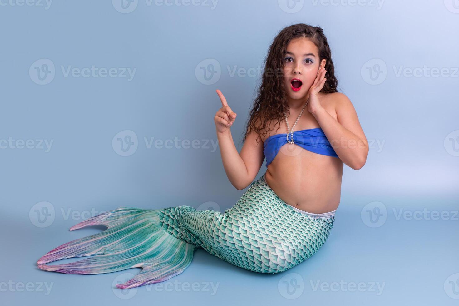A surprised girl in a mermaid costume sits on a blue background photo
