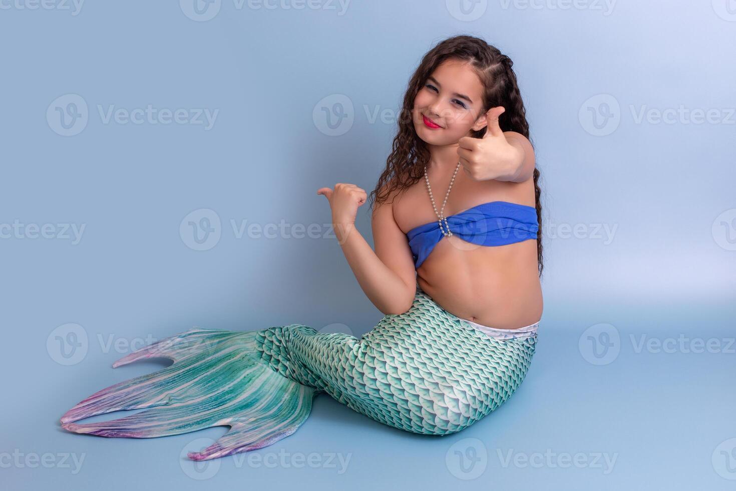 A happy girl in a mermaid costume sitting on a blue background showing thumbs up. photo