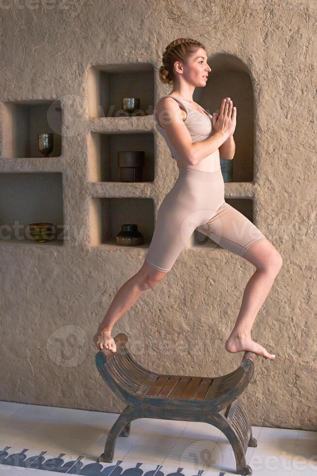 A slender beautiful woman in a beige top and leggings stands in a yoga pose on a chair photo
