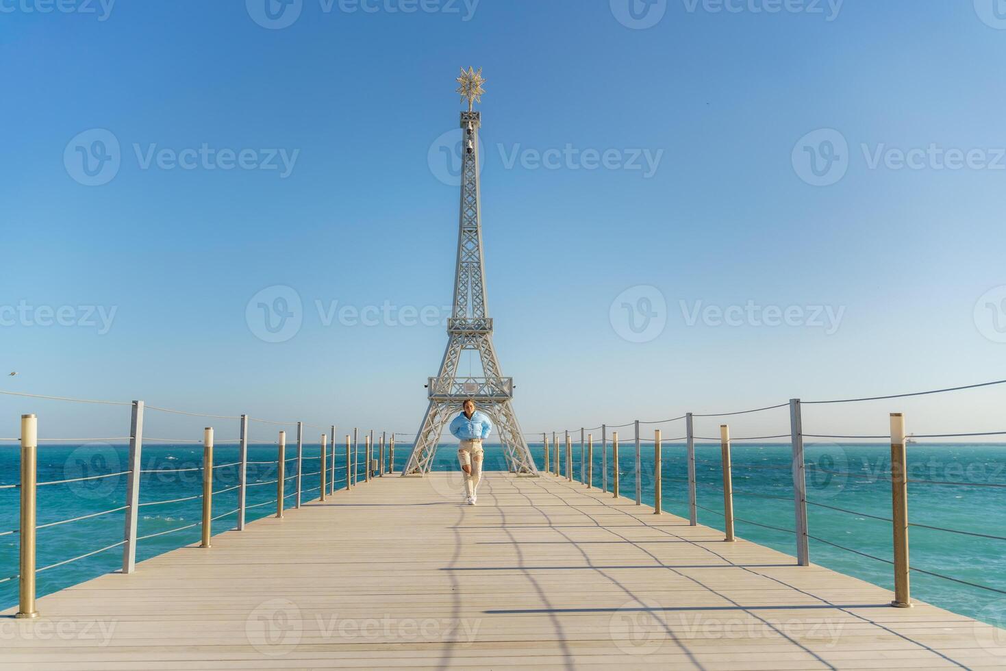 Large model of the Eiffel Tower on the beach. A woman walks along the pier towards the tower, wearing a blue jacket and white jeans. photo
