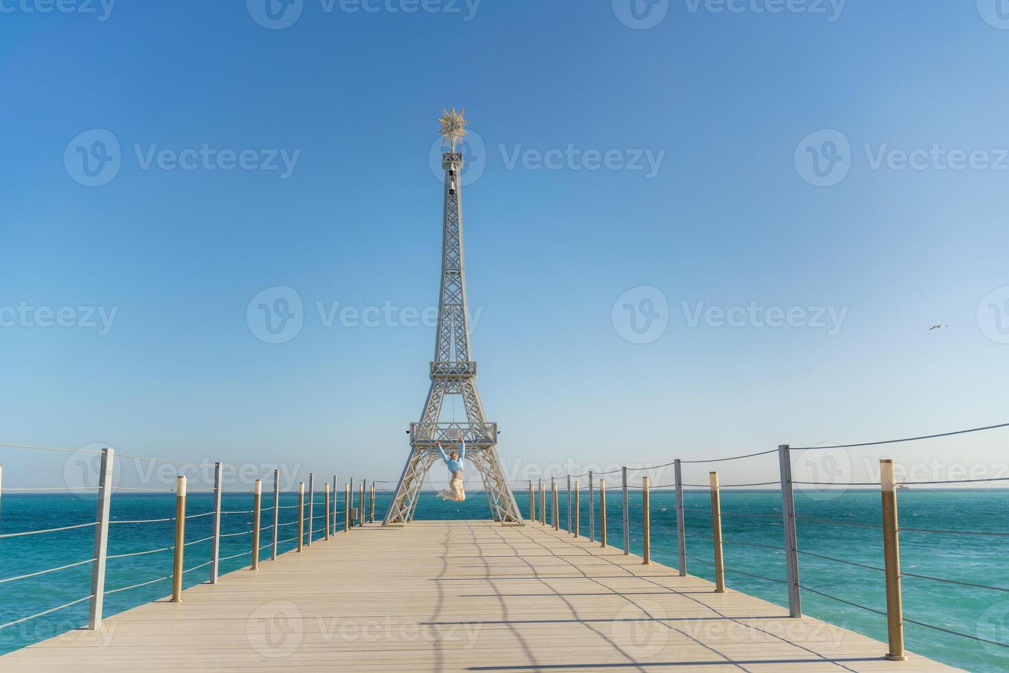 Large model of the Eiffel Tower on the beach. A woman walks along the pier towards the tower, wearing a blue jacket and white jeans. photo