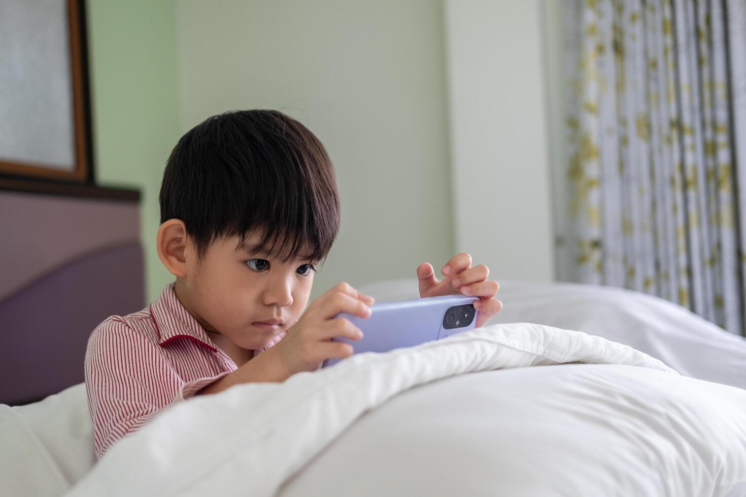 A little Asian boy lies on the bed playing with his phone. photo