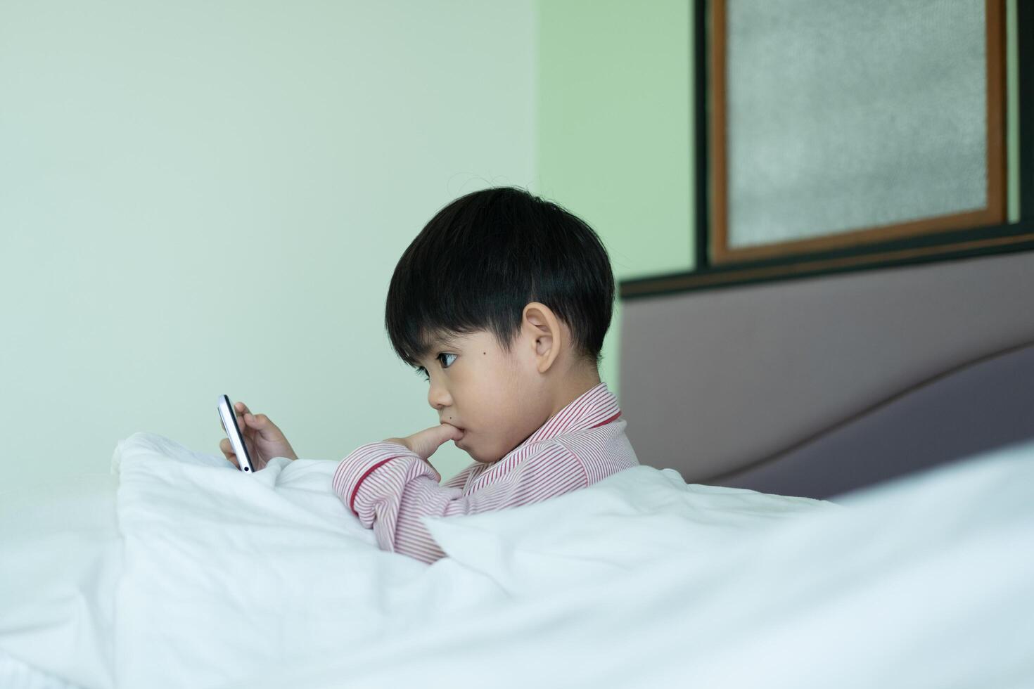 A little Asian boy lies on the bed playing with his phone. photo