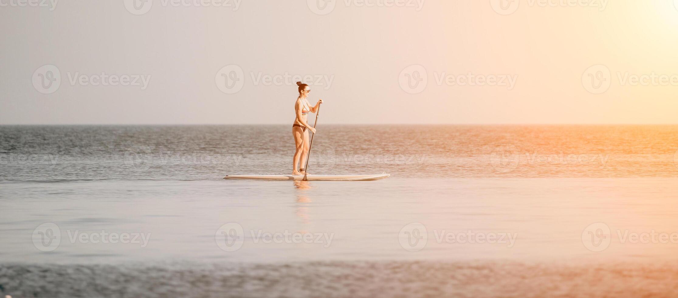 Sea woman sup. Silhouette of happy middle aged tanned woman in rainbow bikini, surfing on SUP board, confident paddling through water surface. Idyllic sunset. Active lifestyle at sea or river. photo