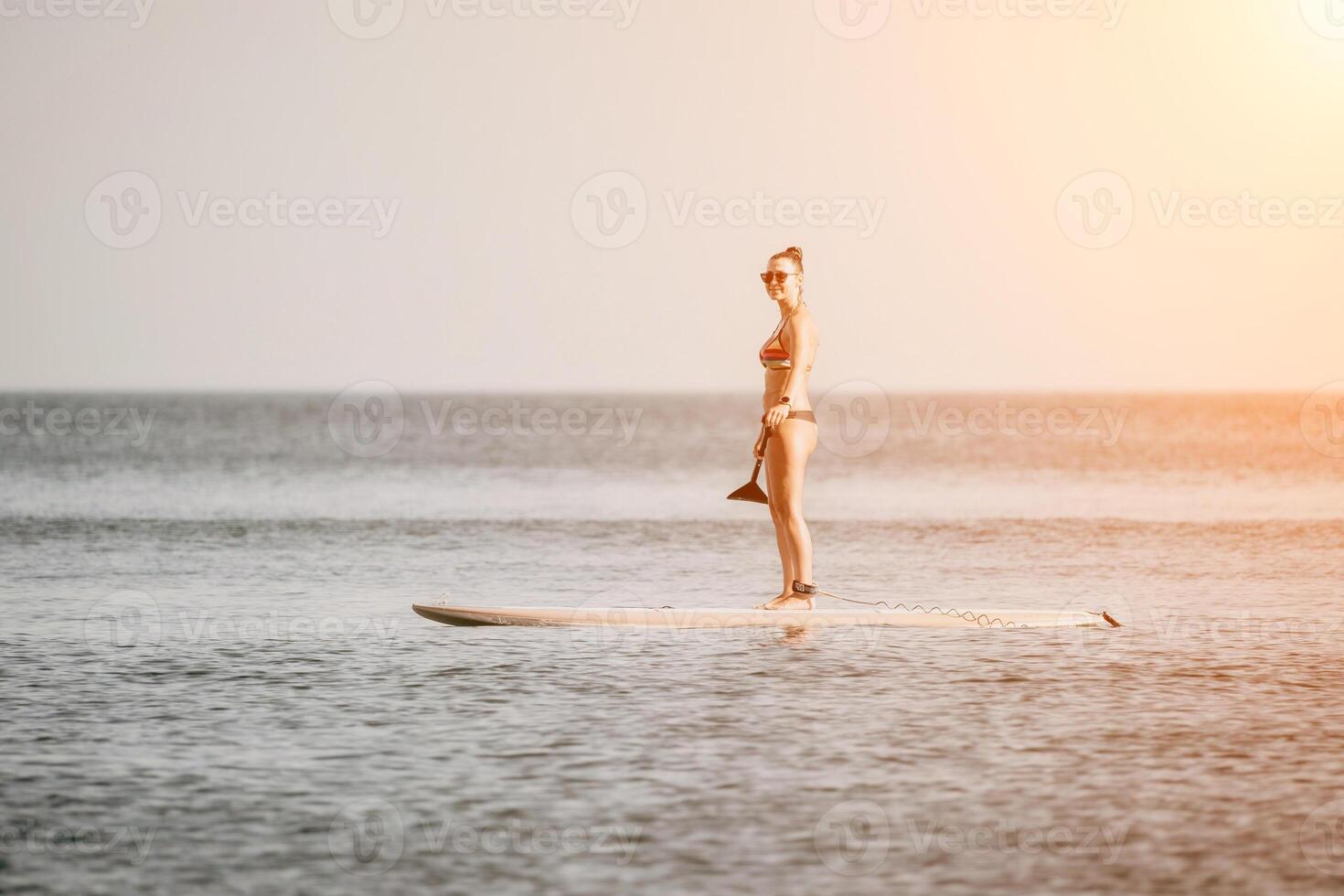 Sea woman sup. Happy woman in a rainbow-coloured bikini stands on SUP board. She is paddling through the calm water, creating ripples along the way. Idyllic sunset. Active lifestyle at sea or river. photo