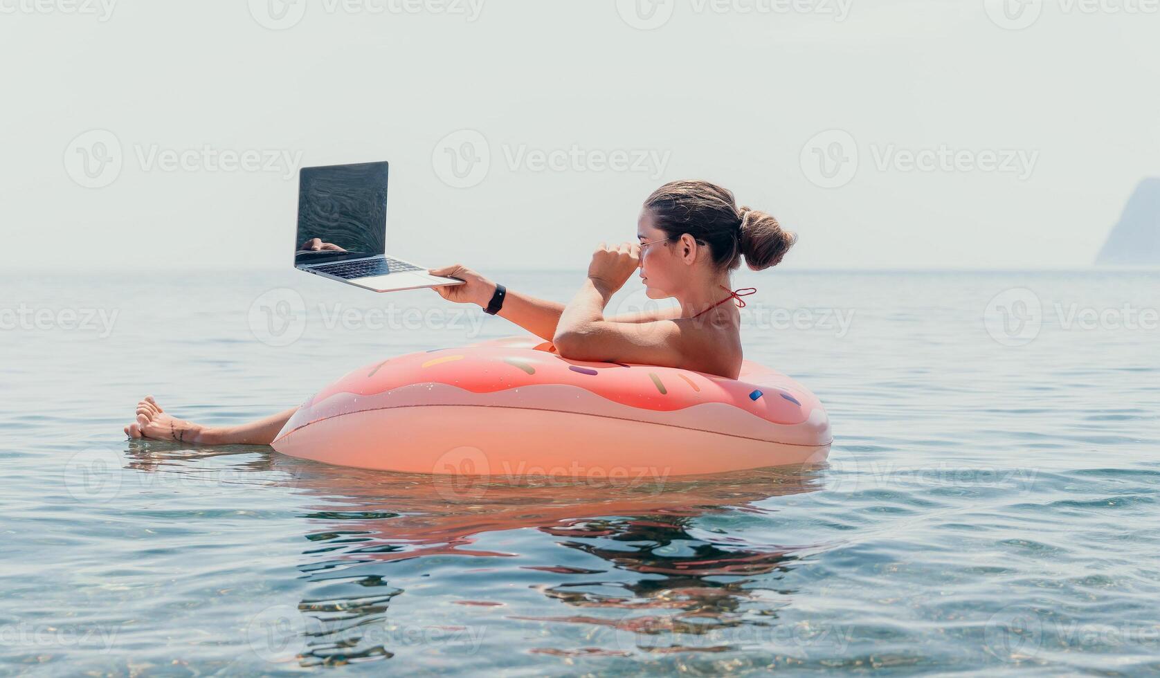 Woman freelancer works on laptop swimming in sea on pink inflatable ring. Happy tourist floating on inflatable donut and working on laptop computer in calm ocean. Freelance, remote working anywhere photo