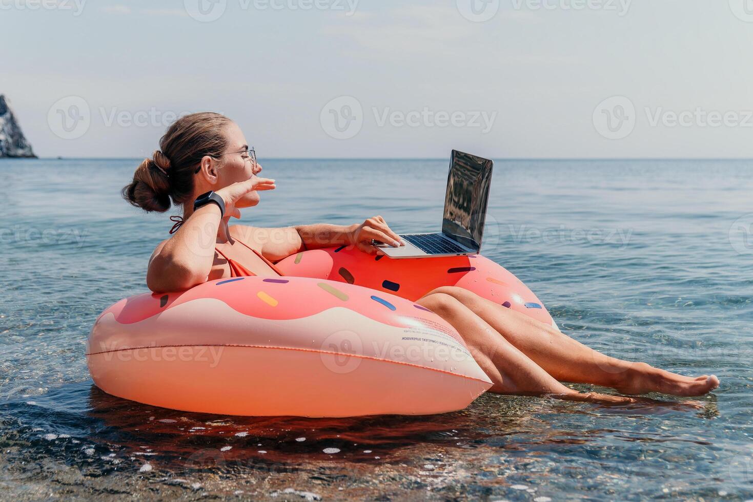 Woman freelancer works on laptop swimming in sea on pink inflatable ring. Happy tourist floating on inflatable donut and working on laptop computer in calm ocean. Freelance, remote working anywhere photo
