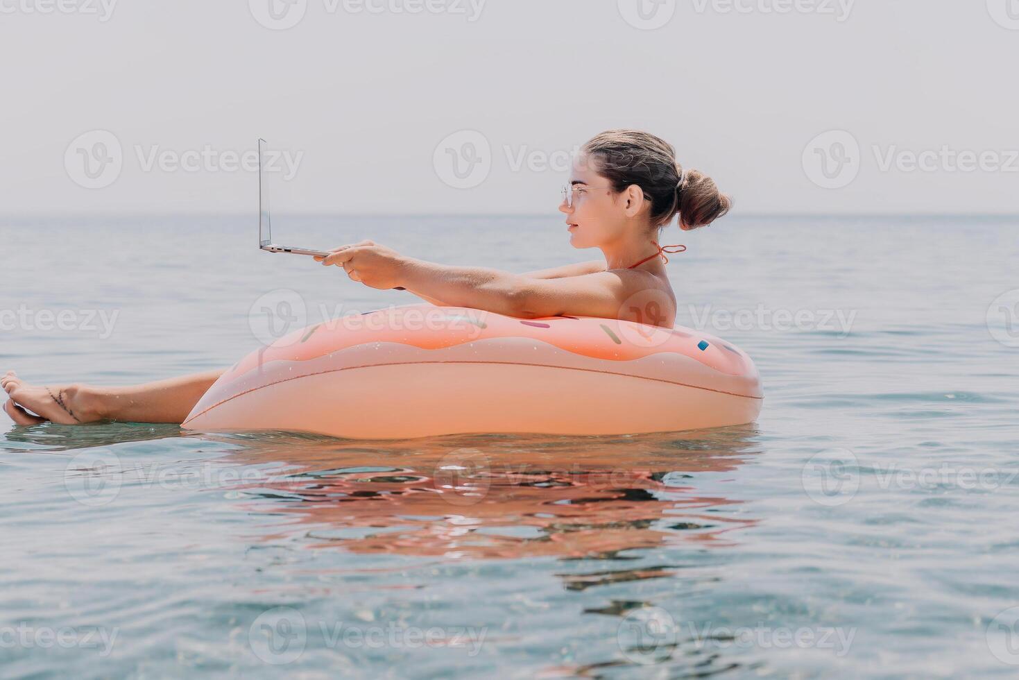 Woman freelancer works on laptop swimming in sea on pink inflatable ring. Happy tourist floating on inflatable donut and working on laptop computer in calm ocean. Freelance, remote working anywhere photo