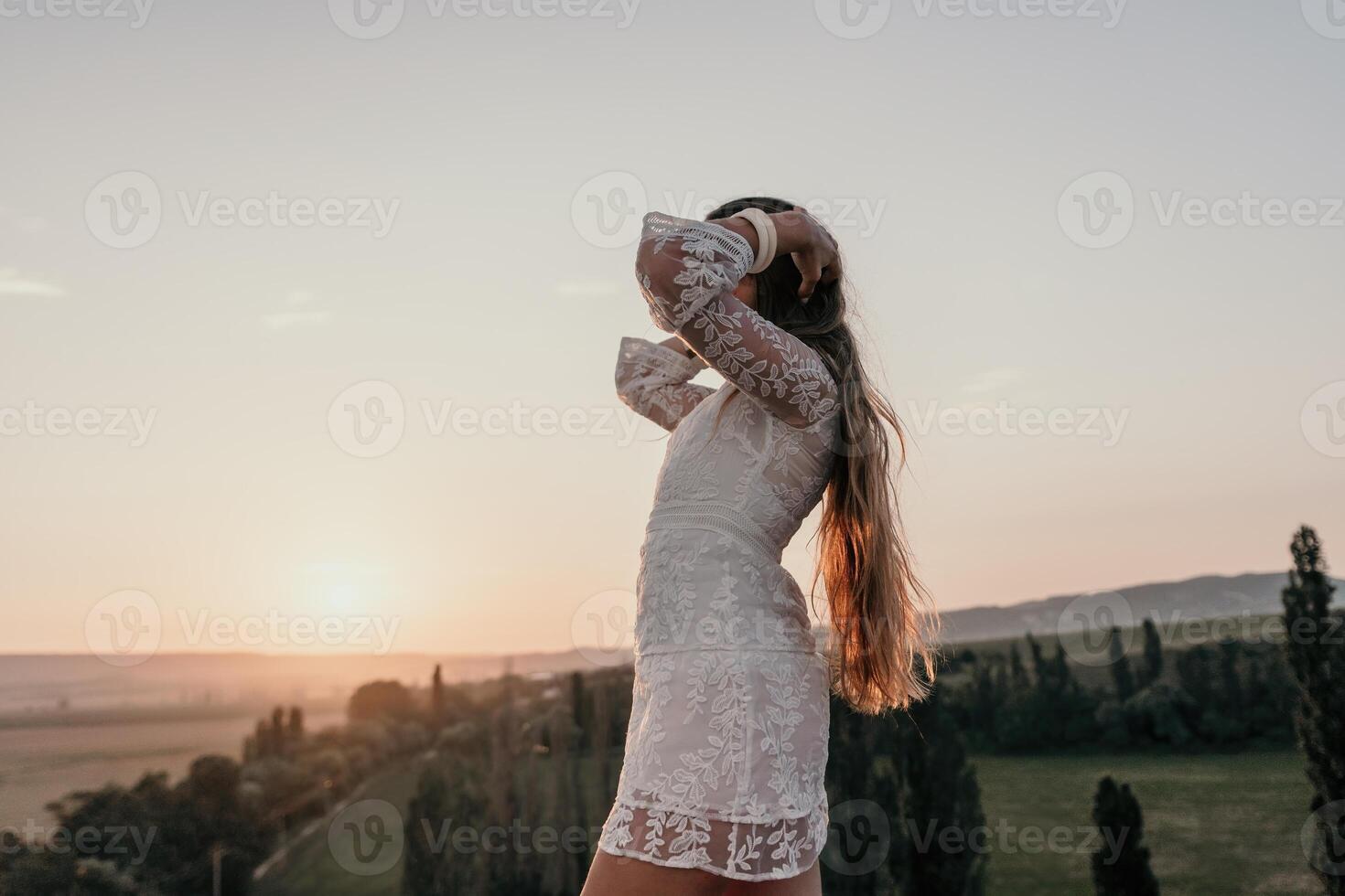 Happy woman in white boho dress on sunset in mountains. Romantic woman with long hair standing with her back on the sunset in nature in summer with open hands. Silhouette. Nature. Sunset. photo