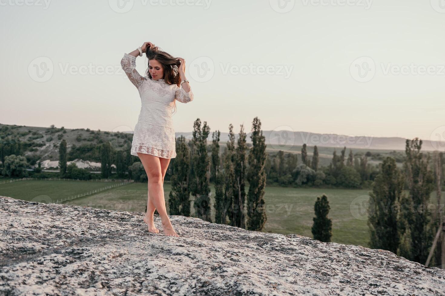 Happy woman in white boho dress on sunset in mountains. Romantic woman with long hair standing with her back on the sunset in nature in summer with open hands. Silhouette. Nature. Sunset. photo