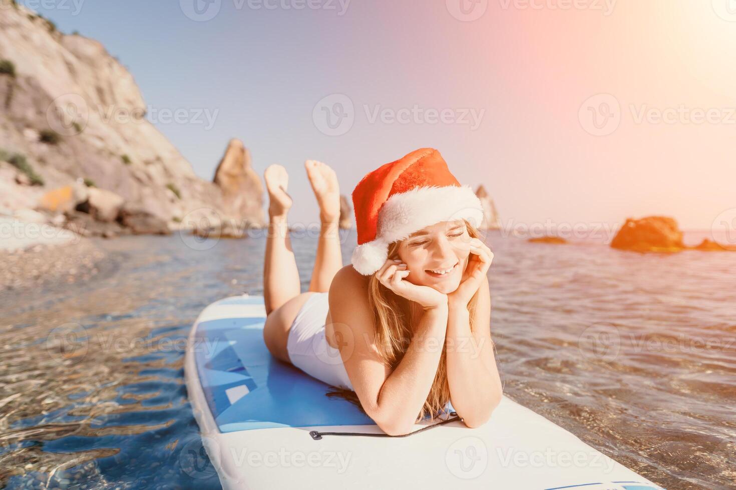Woman sea sup. Close up portrait of happy young caucasian woman with long hair in Santa hat looking at camera and smiling. Cute woman portrait in a white bikini posing on sup board in the sea photo