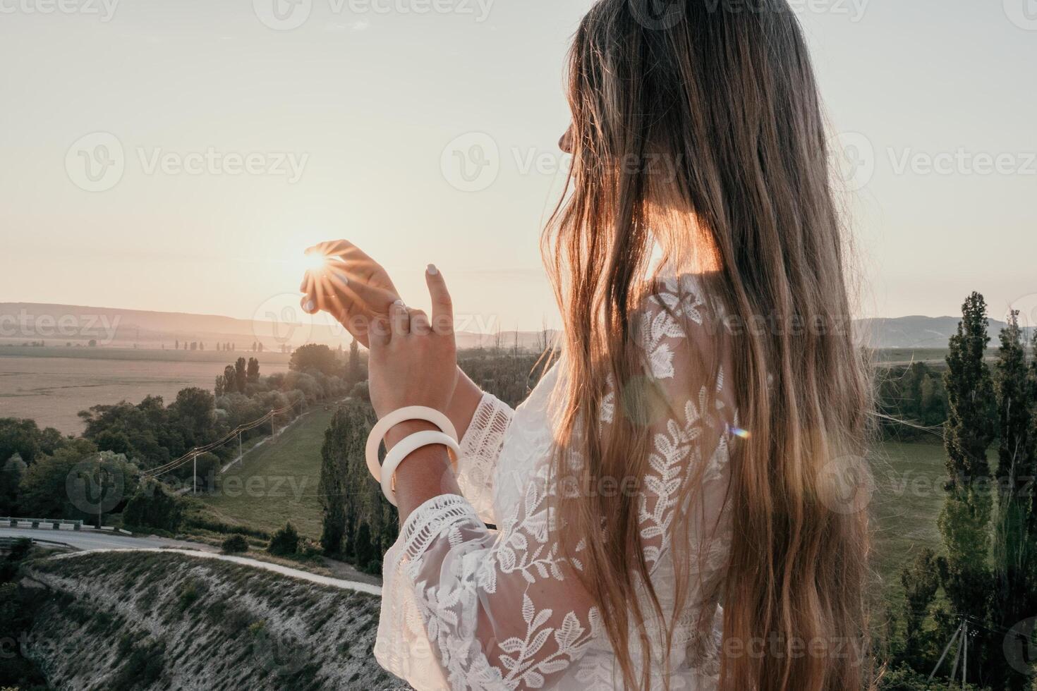 Happy woman in white boho dress on sunset in mountains. Romantic woman with long hair standing with her back on the sunset in nature in summer with open hands. Silhouette. Nature. Sunset. photo