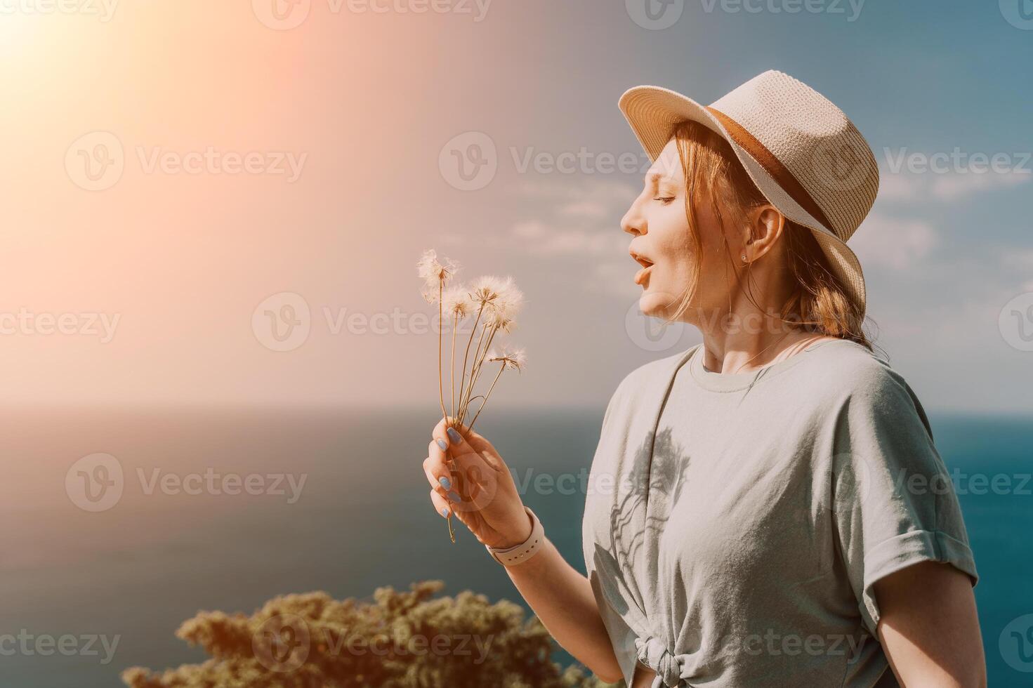 Woman travel sea. Happy tourist in hat standing on a beach and holding a bouquet of dandelion flowers. She is blowing the petals into the air, creating a beautiful and serene scene. photo