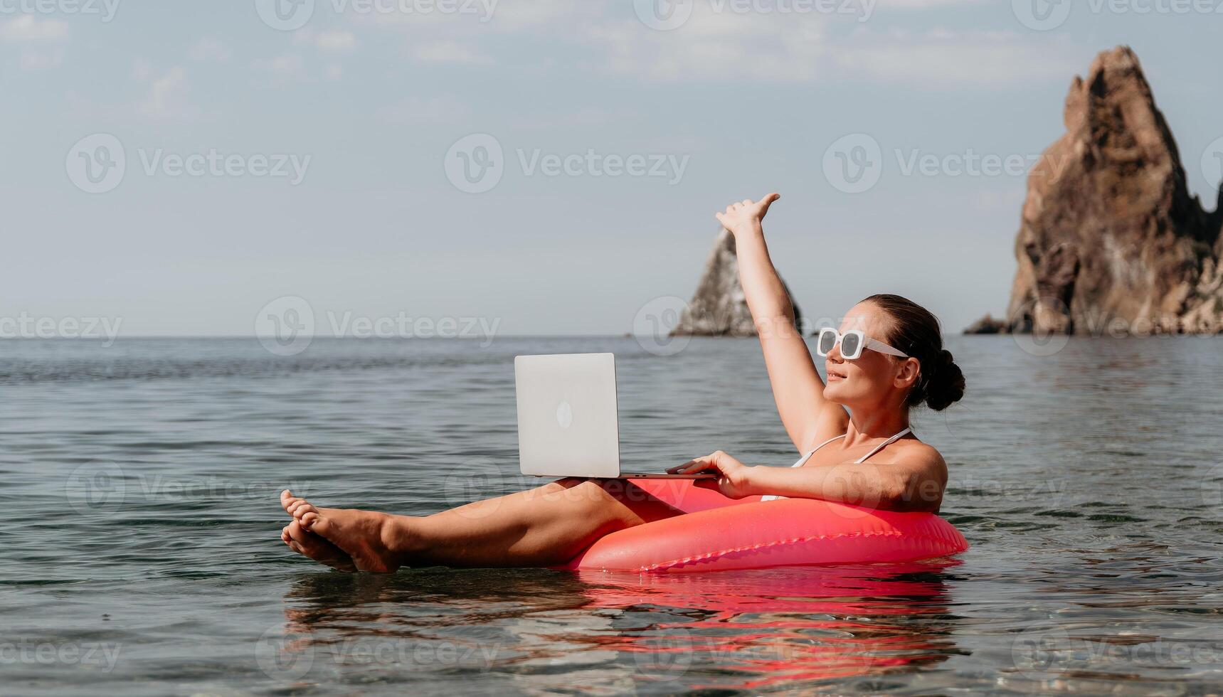 Woman freelancer works on laptop swimming in sea on pink inflatable ring. Happy tourist in sunglasses floating on inflatable donut and working on laptop computer in calm ocean. Remote working anywhere photo